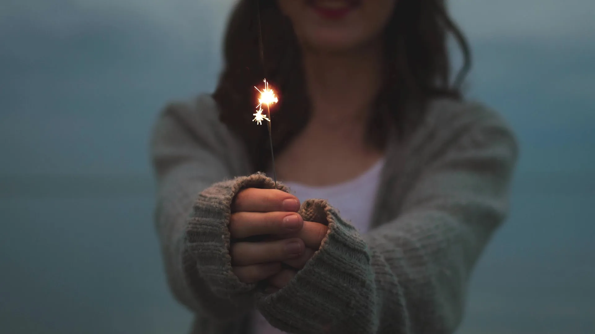 A person in a gray sweater holds a lit sparkler toward the camera.