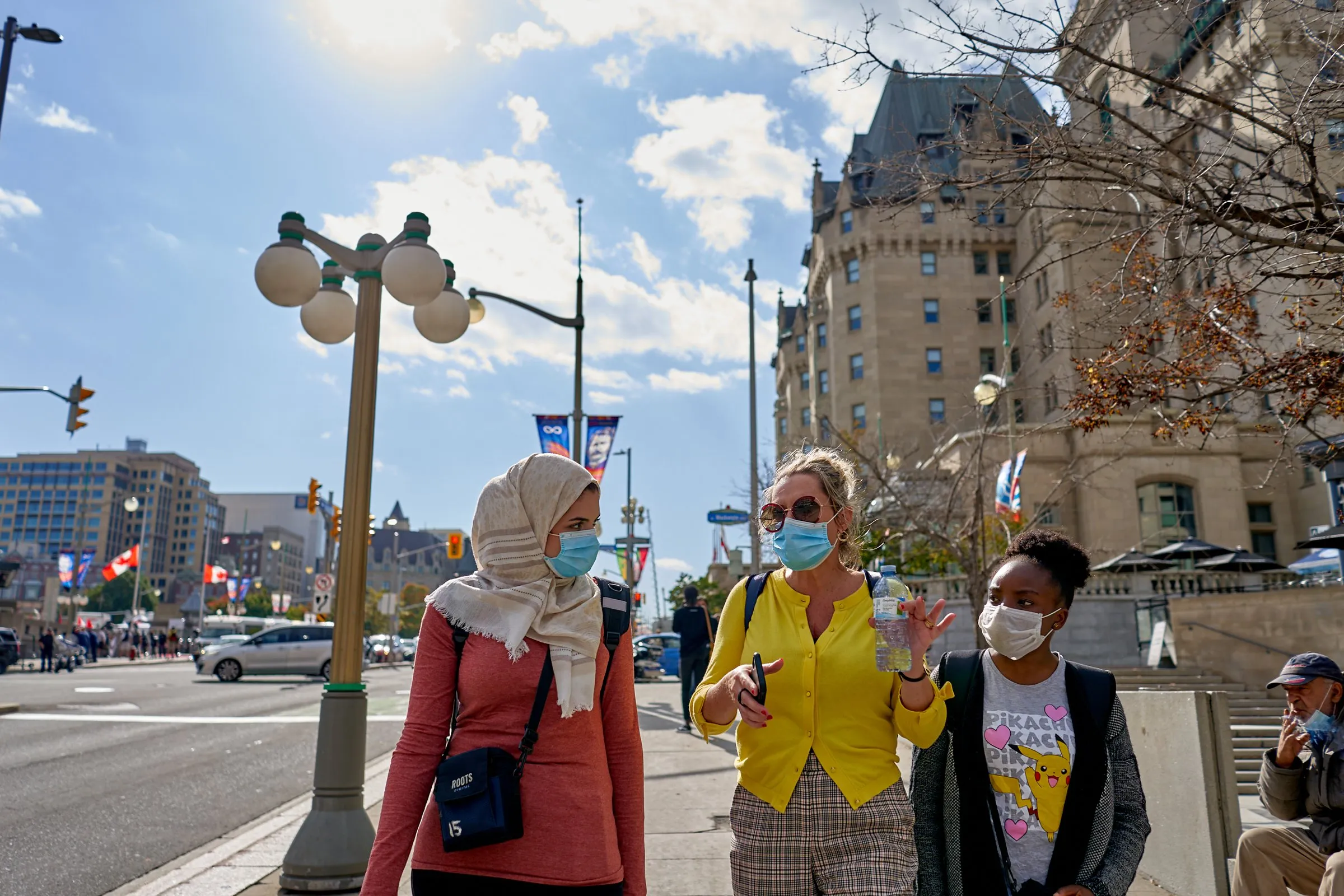 Three masked women walk along a city sidewalk beside a busy street.