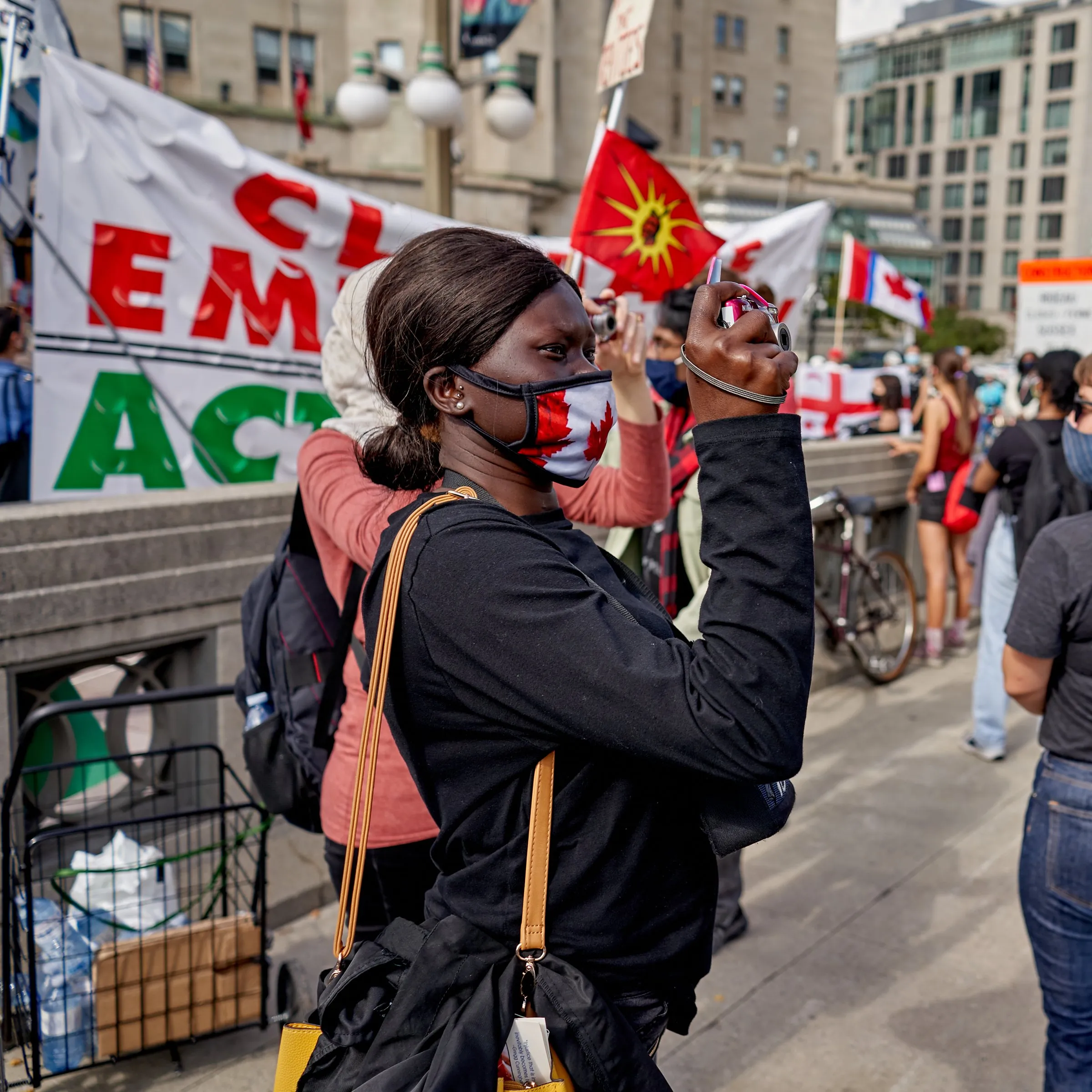 A masked woman holds up a phone at a street protest with flags and banners.