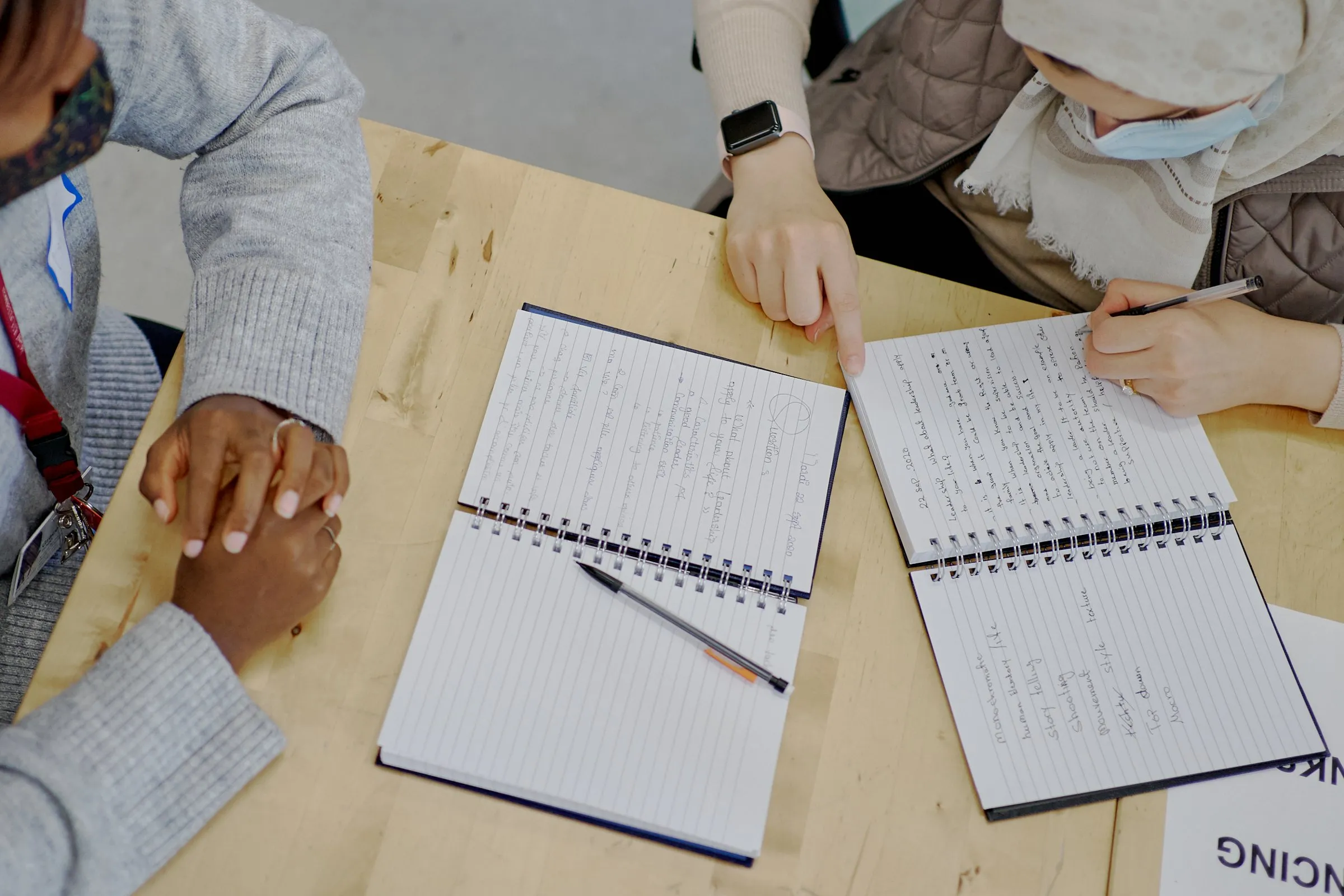 People gathered around a table with open notebooks, one writing and another pointing at a page.