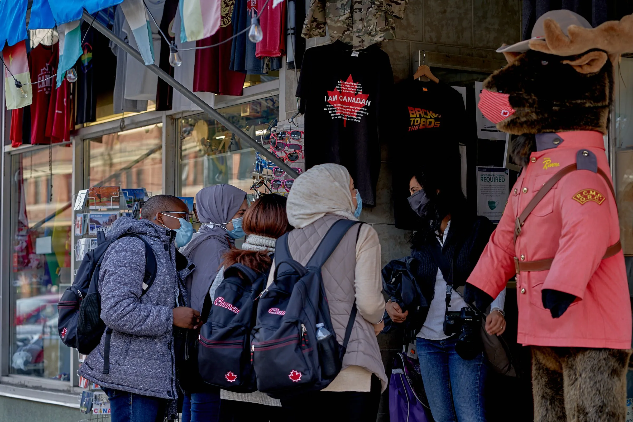 Masked shoppers browse a clothing stall with hanging shirts beside a figure in a pink uniform and furry animal head.