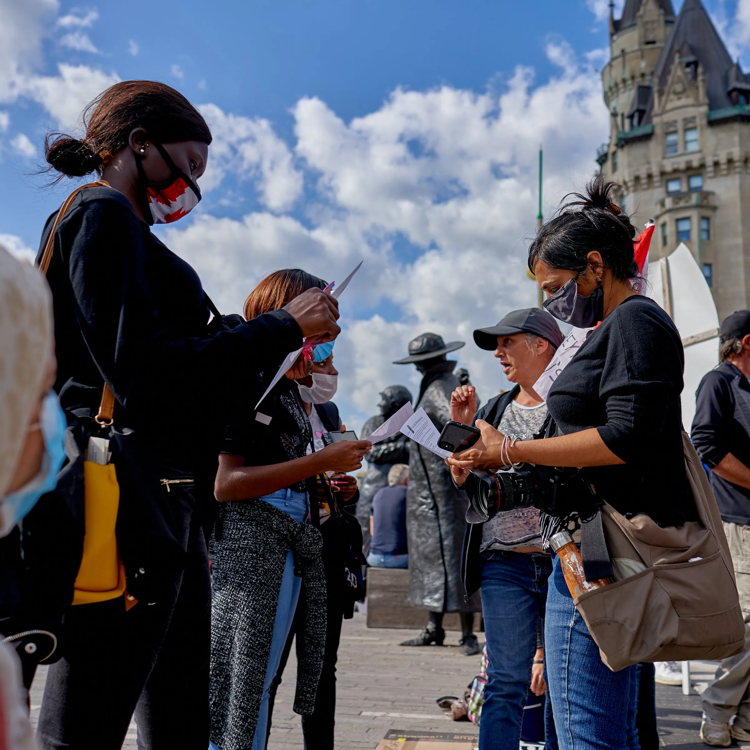 People talk and exchange papers in a city square beneath a partly cloudy sky.