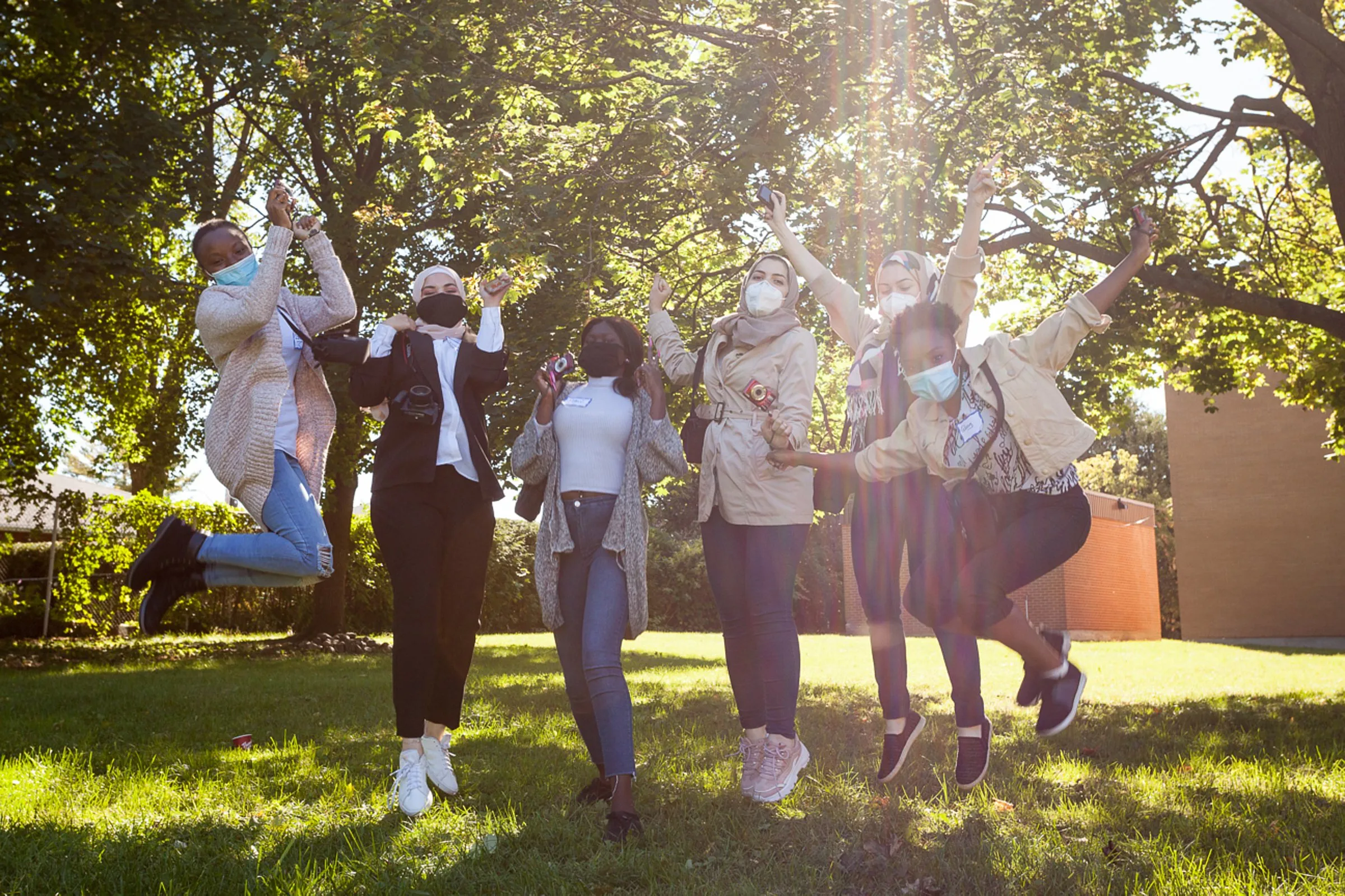 A group of masked people celebrate outdoors beneath trees in a sunny park.
