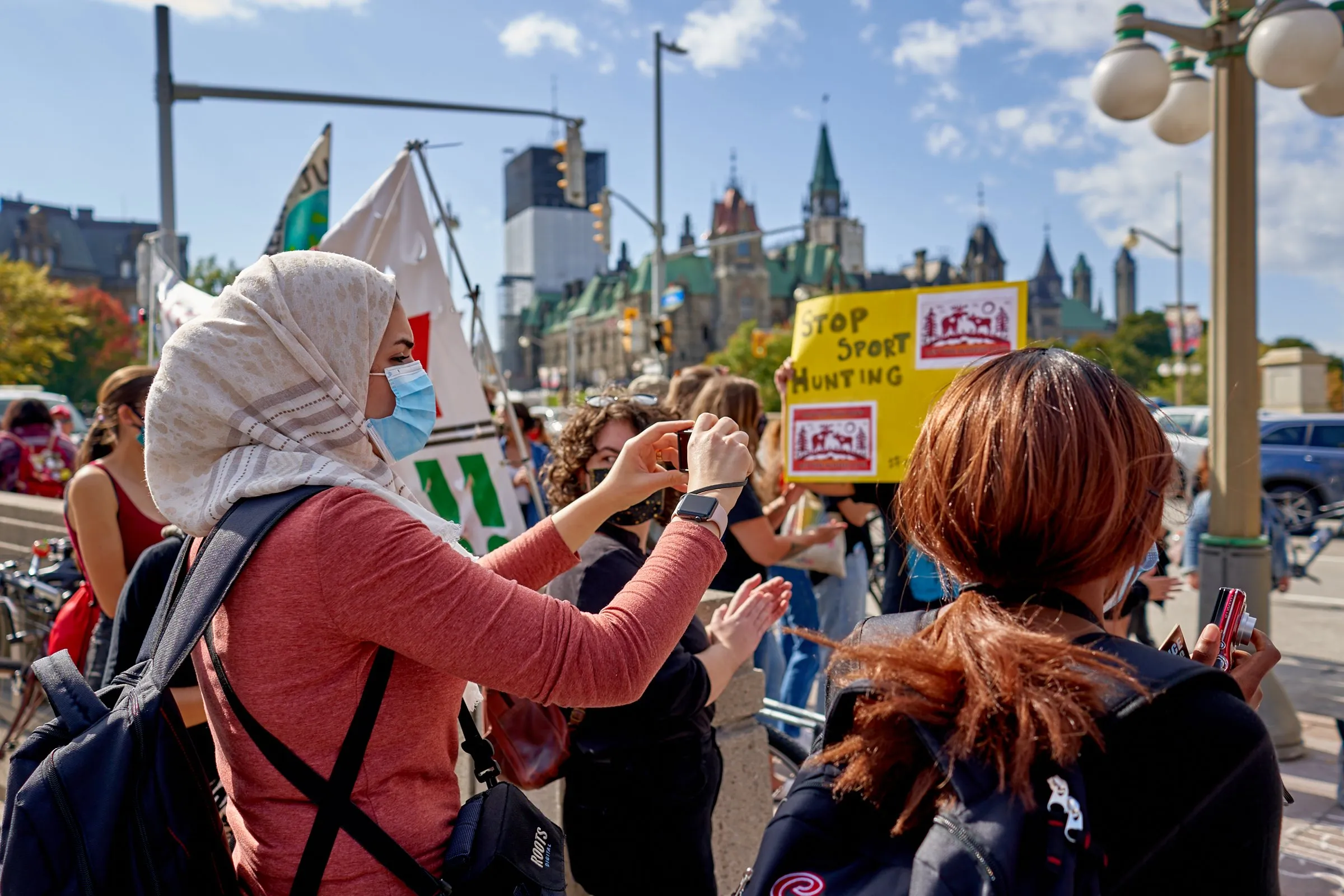 People gather at a street protest; a woman photographs signs, including one reading “Stop Sport Hunting.”