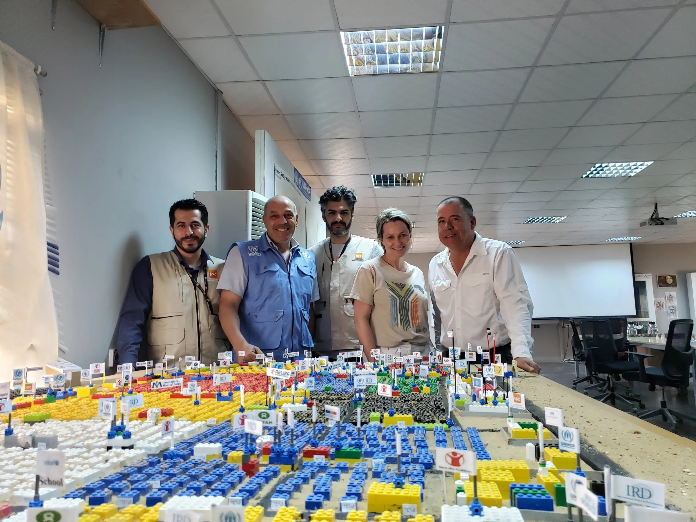 Five adults stand behind a large tabletop display of colorful blocks and small signs in a classroom.