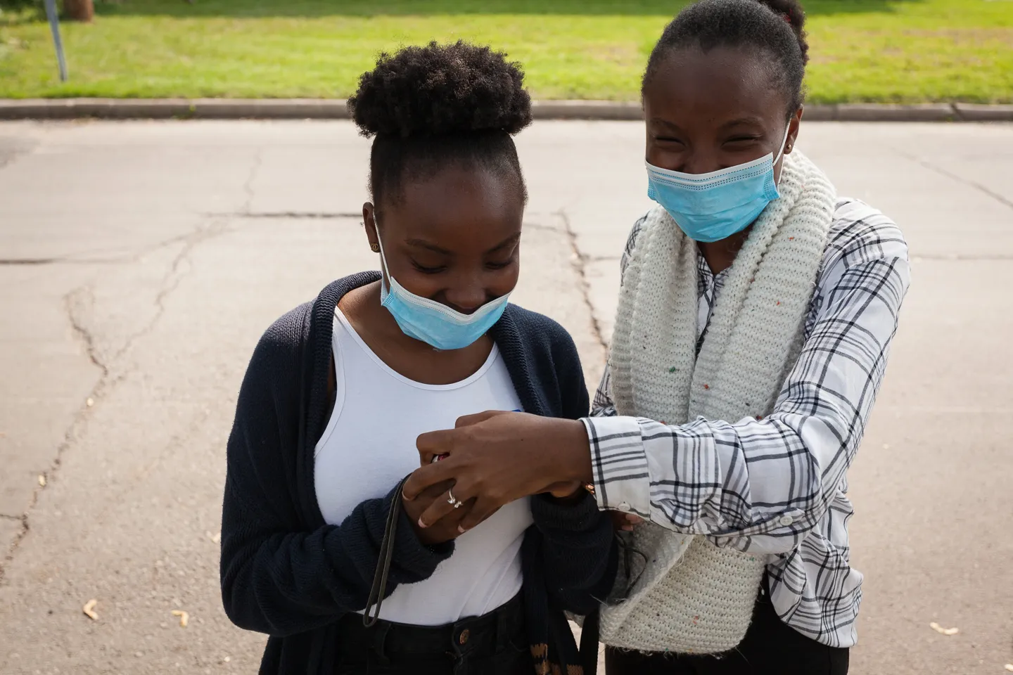 Two people wearing face masks outdoors; one helps the other adjust a bag strap.