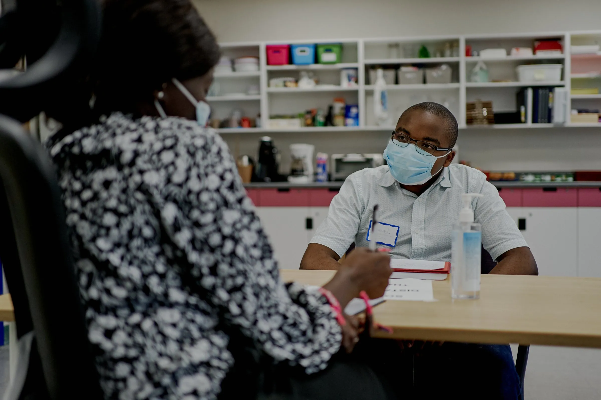 A masked healthcare worker speaks with a woman across a table in a clinic.