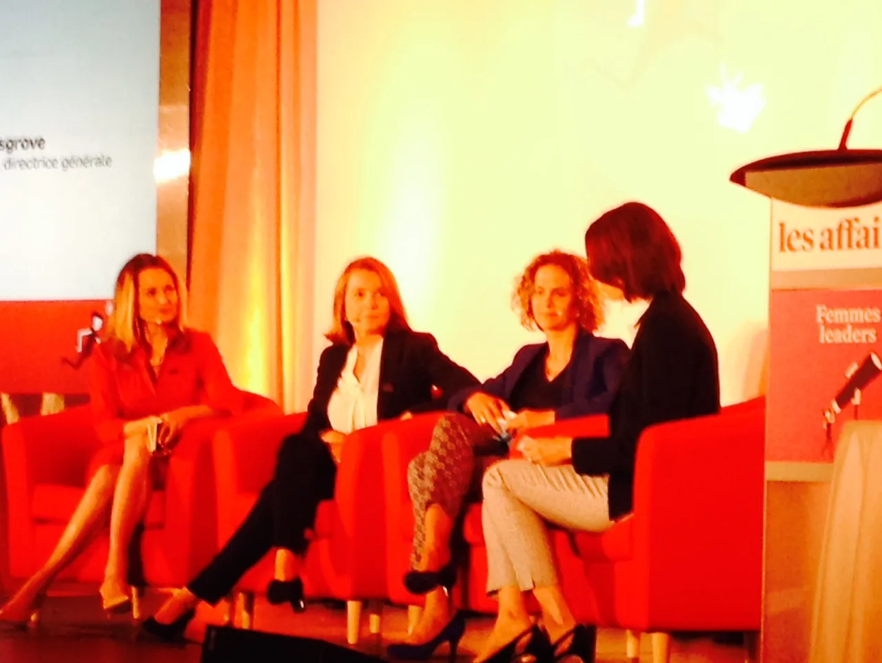 Four women seated in red chairs during a stage discussion beside a podium.
