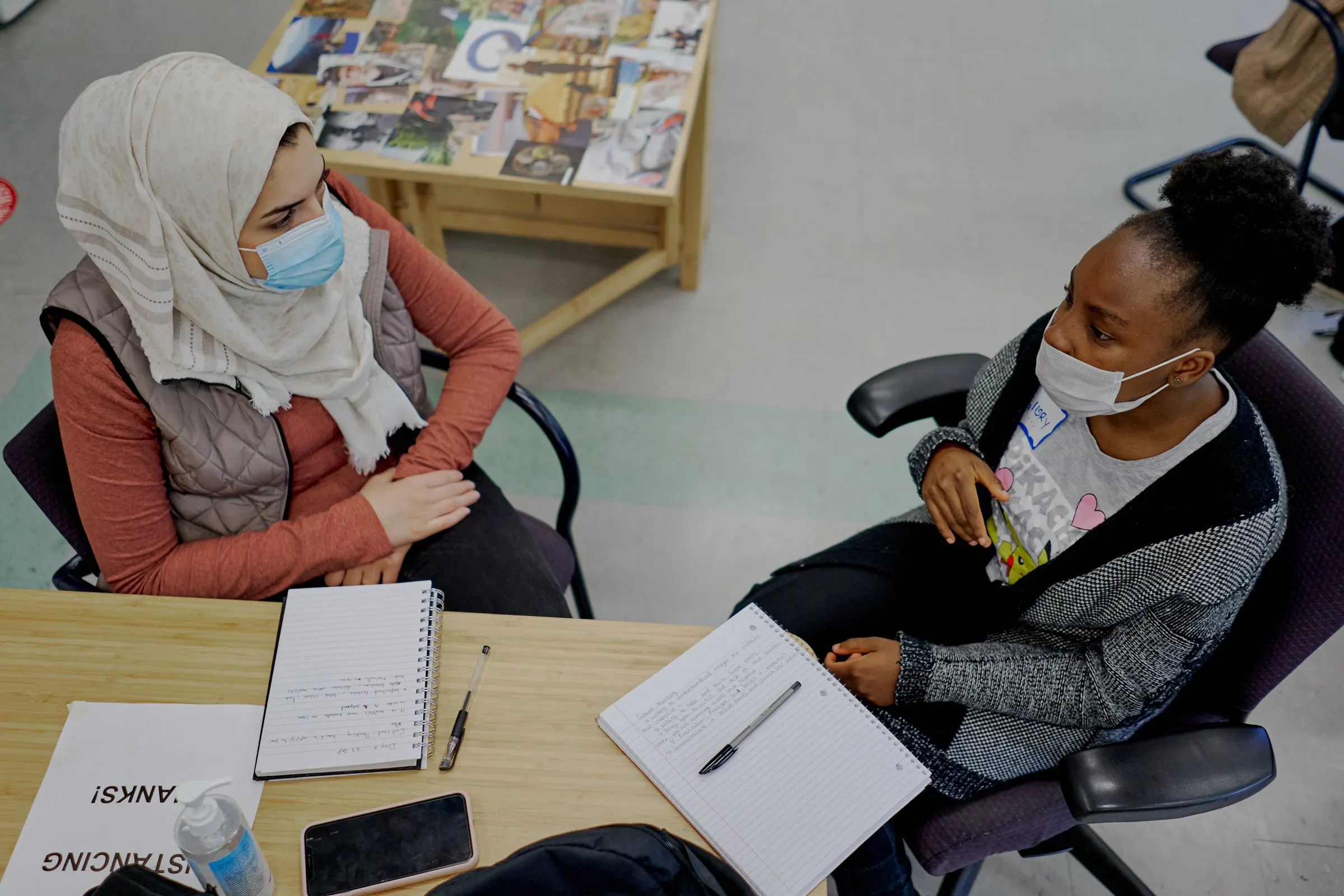 Two masked women sit at a table with open notebooks, talking.