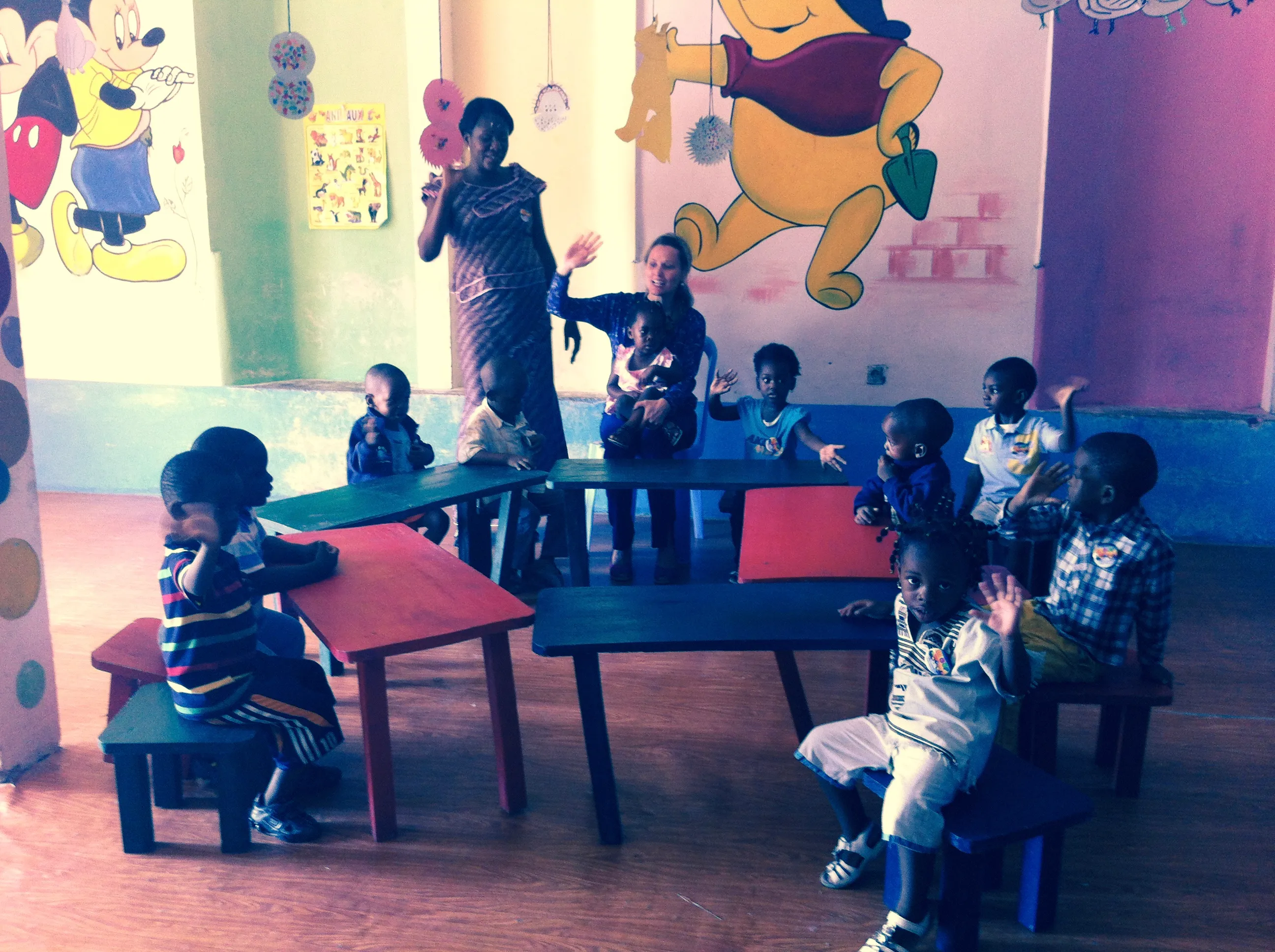 Children sit at low tables in a colorful classroom with cartoon murals, while two adults stand among them.