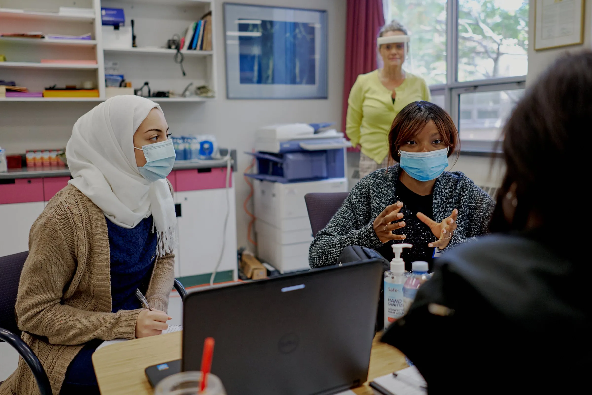Three masked women sit around a laptop in a medical office, with another woman standing behind them.