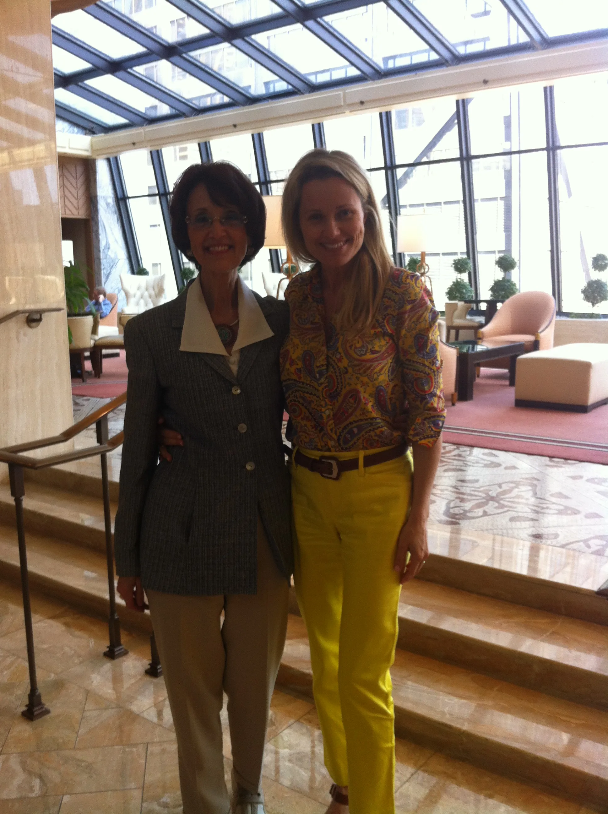 Two women standing together indoors, one wearing a patterned blouse and yellow pants, the other a gray suit.