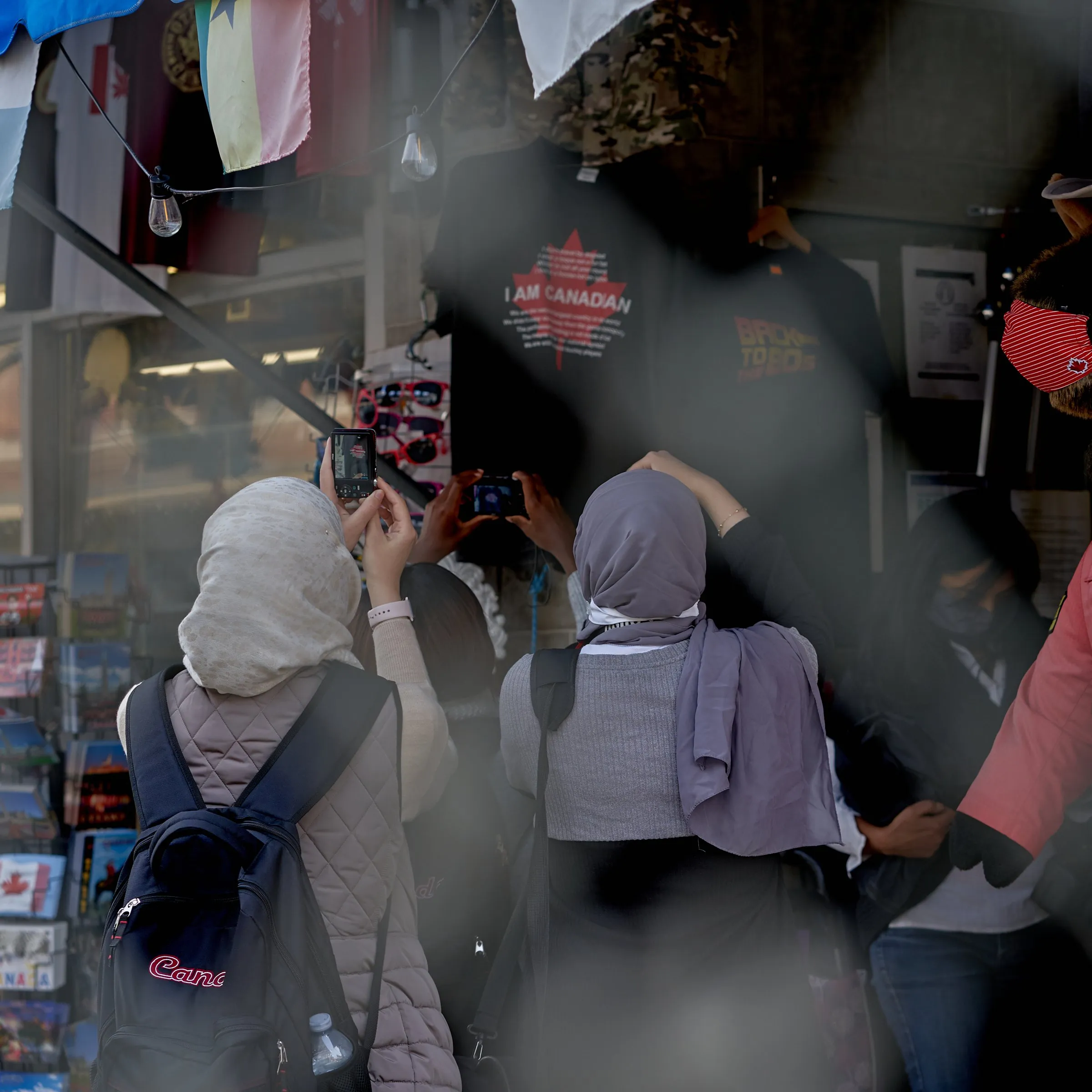 Two people in headscarves photograph a storefront display with an “I AM CANADIAN” shirt.