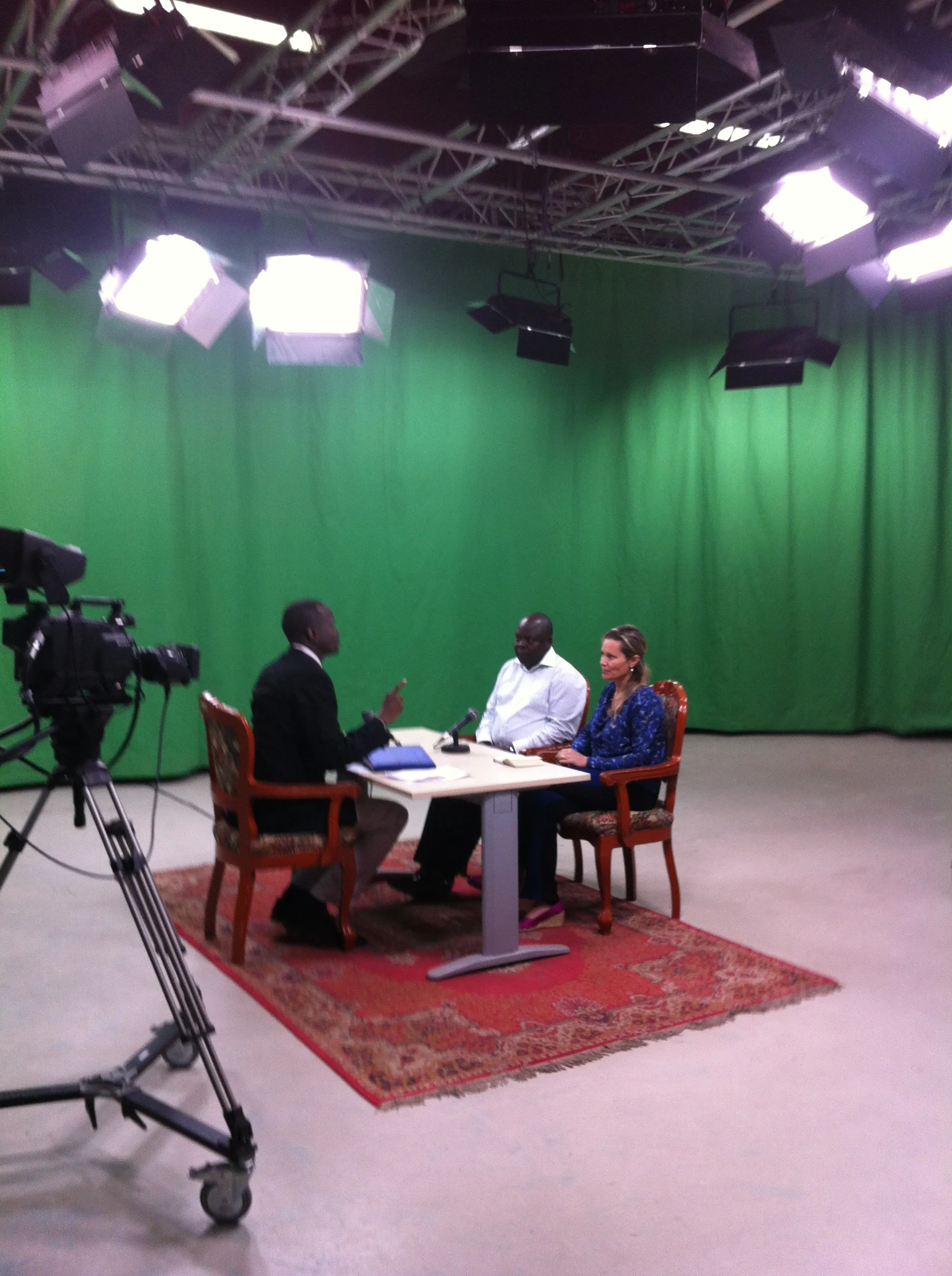 Three people sit around a table in a studio with a green backdrop, lighting rigs, and a video camera.