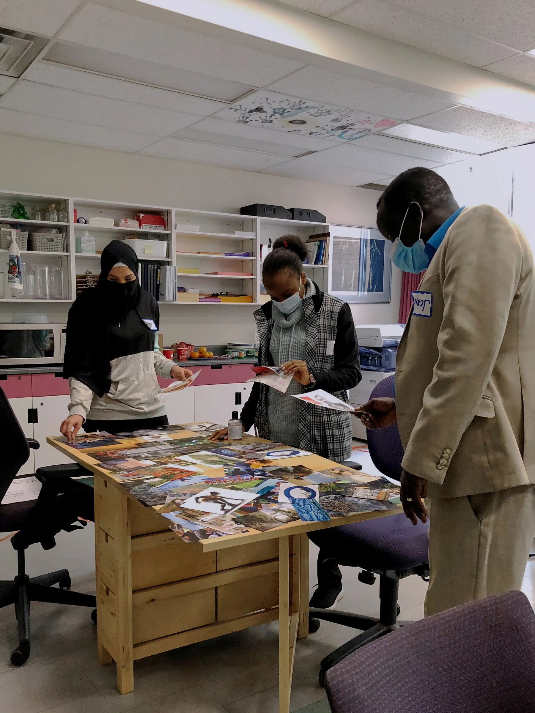 Three masked people examine printed photos and papers on a table in a classroom.