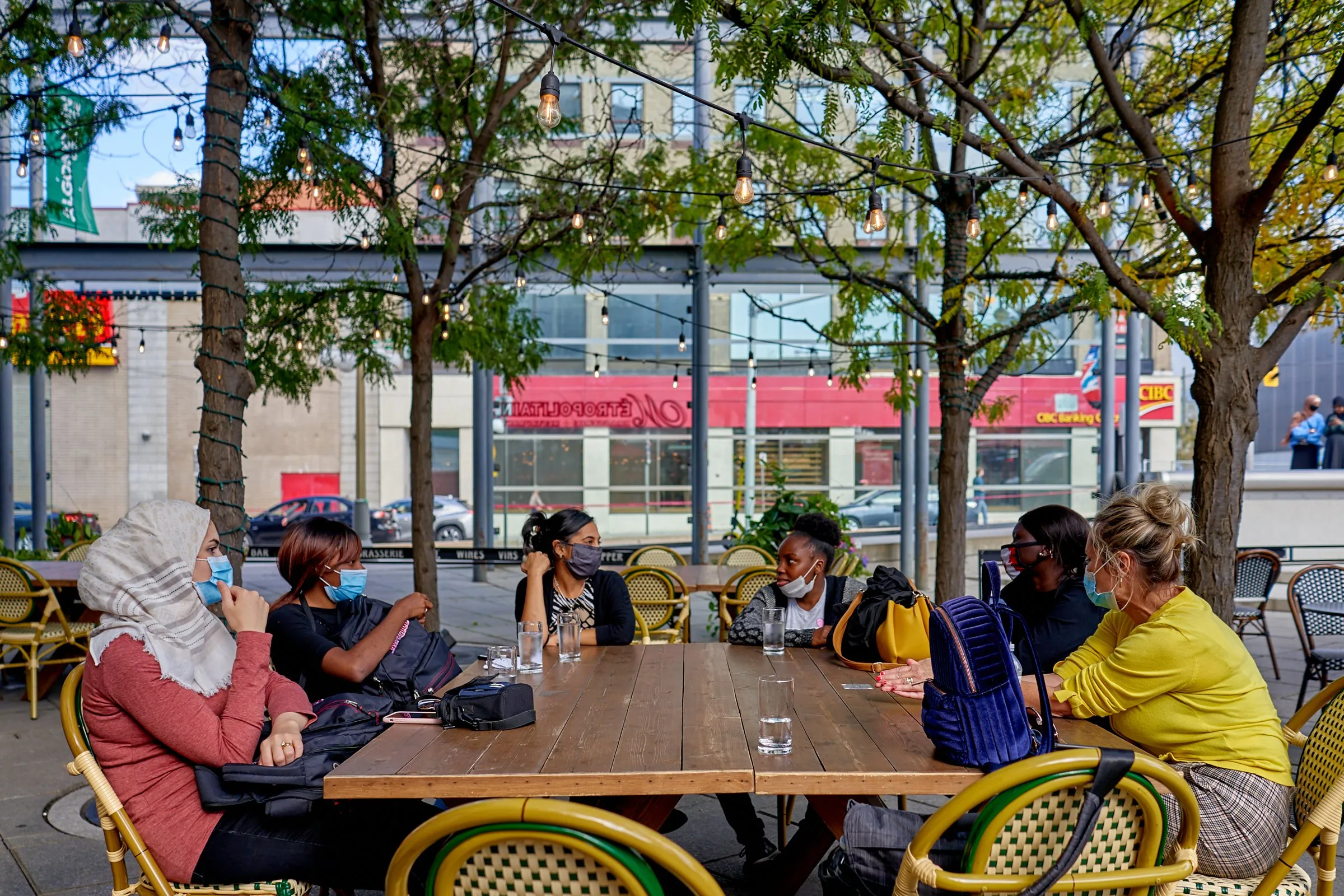 People wearing masks sit around a wooden table at an outdoor café beneath trees.