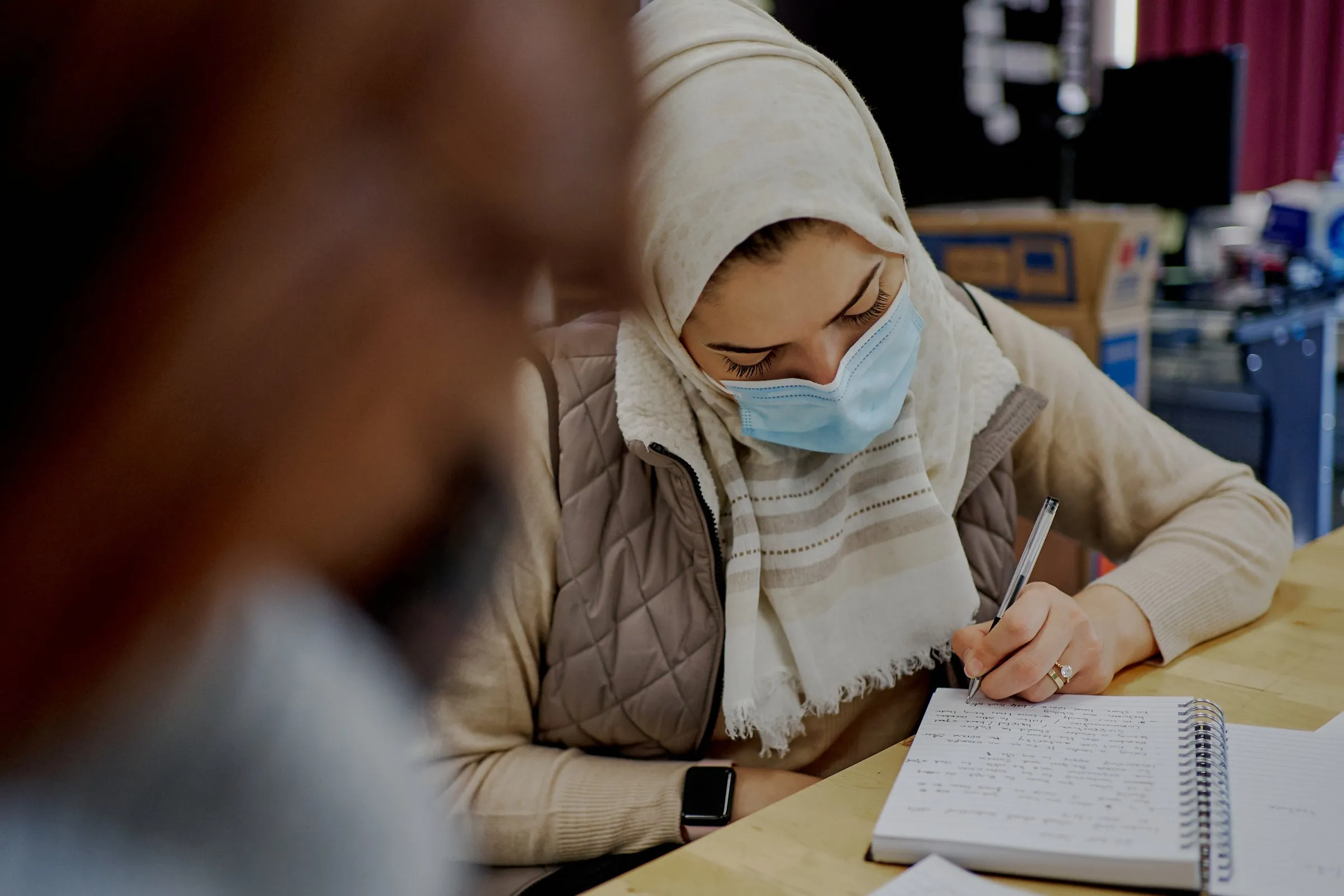 A woman wearing a headscarf and face mask writes in a spiral notebook at a table.