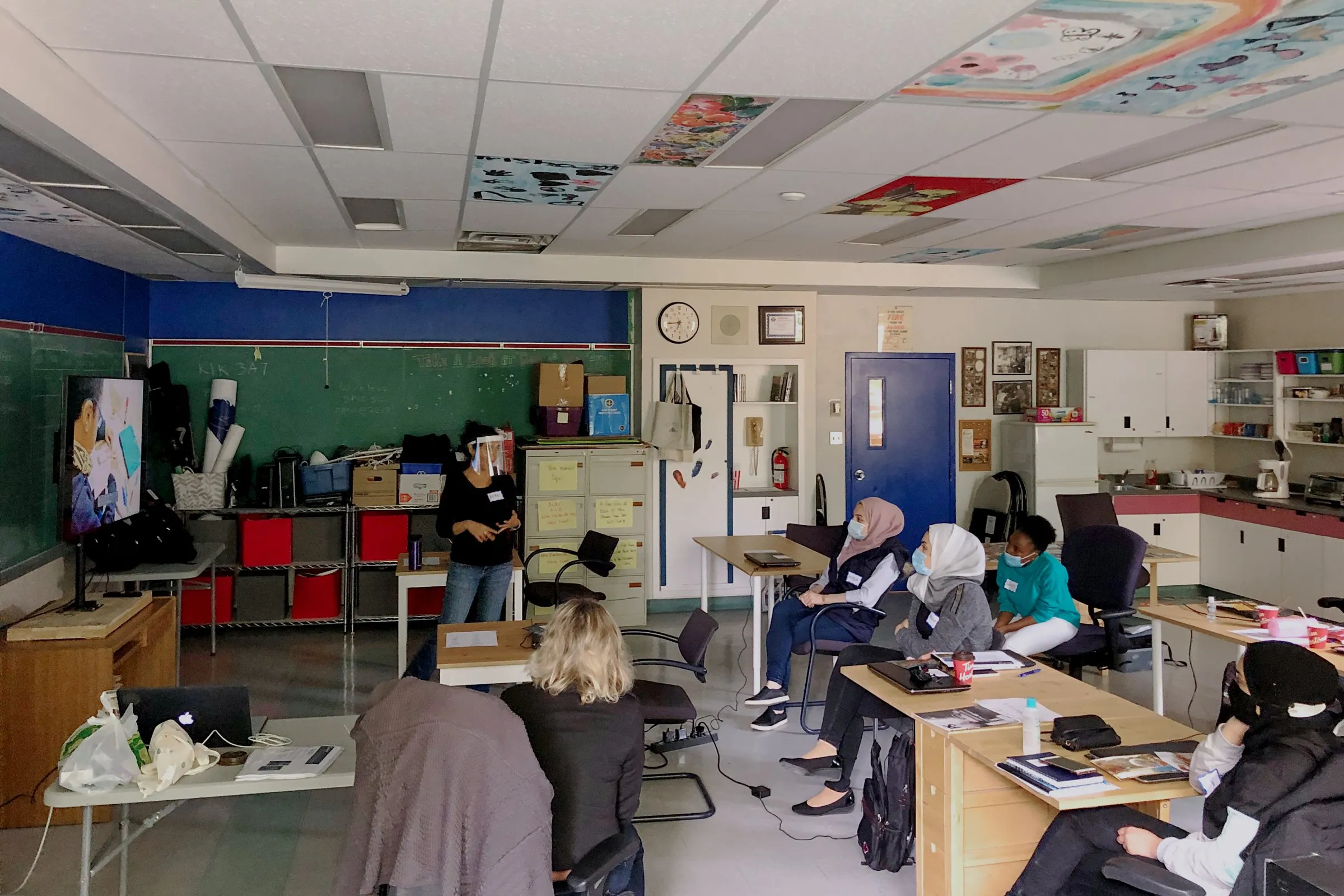 People sit around tables in a classroom as one person stands near a display.