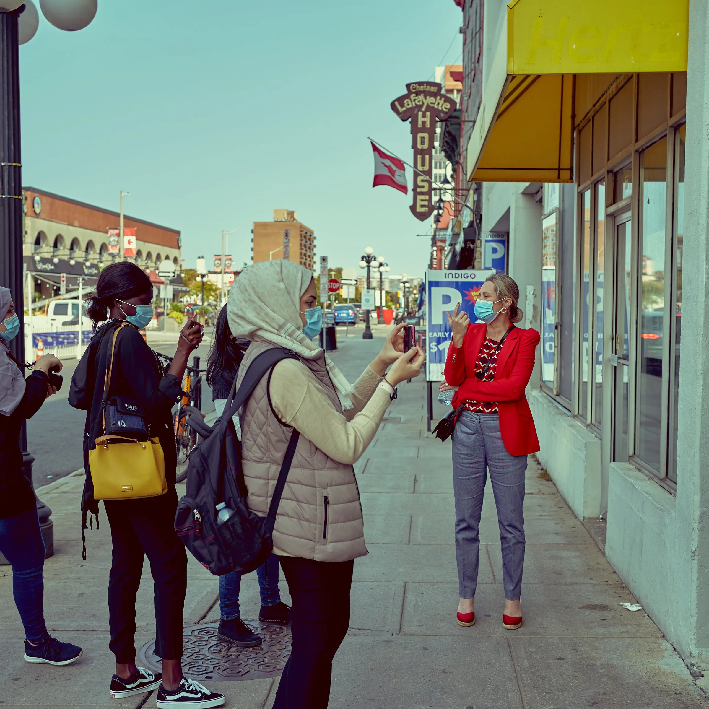 Masked people talk on a city sidewalk; a woman in a red jacket stands facing a woman in a beige coat.