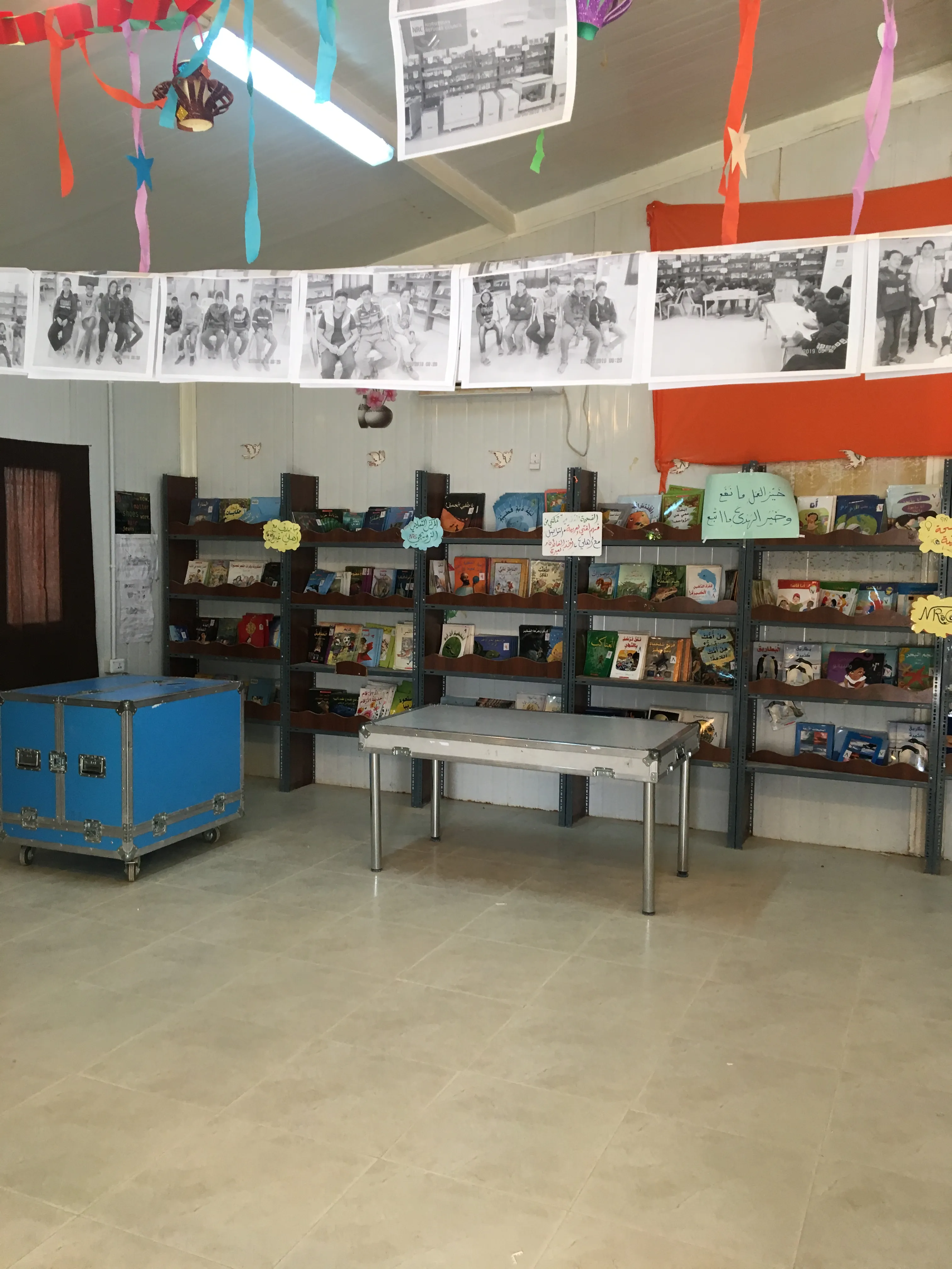 A room with shelves of books and displays, a blue trunk, a table, and hanging black-and-white photo strips.