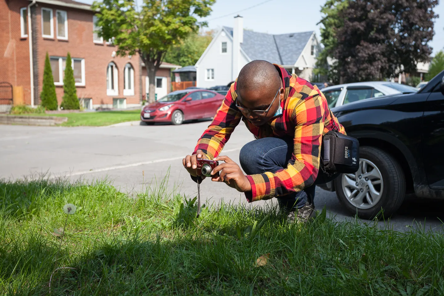 A man crouches by the roadside, photographing grass with a camera.
