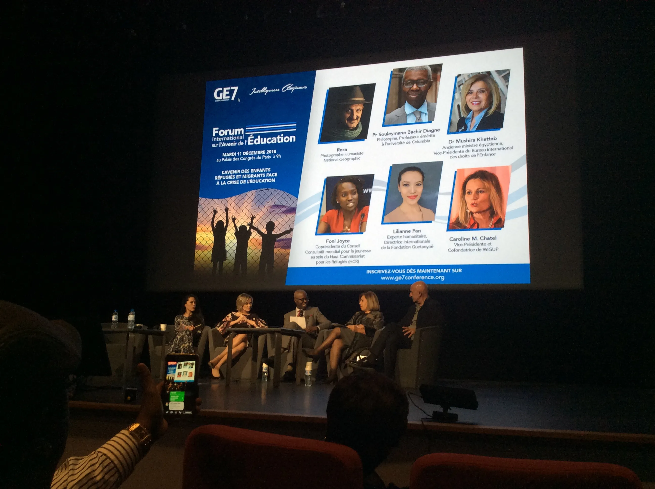 Five people sit on a stage beneath a projected conference poster, while an audience member holds up a phone.