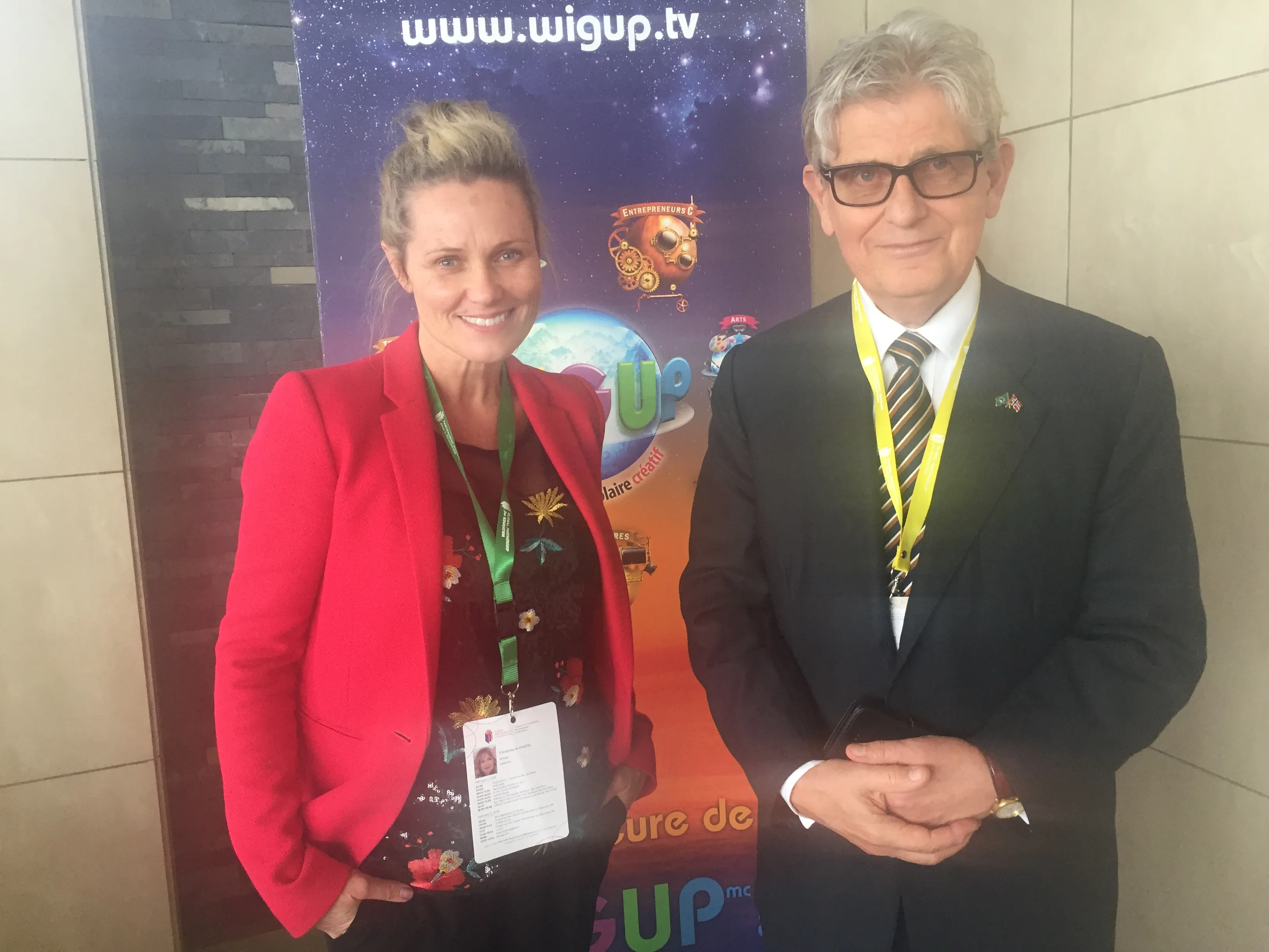 A woman in a red blazer and a man in a dark suit stand in front of a colorful event banner.