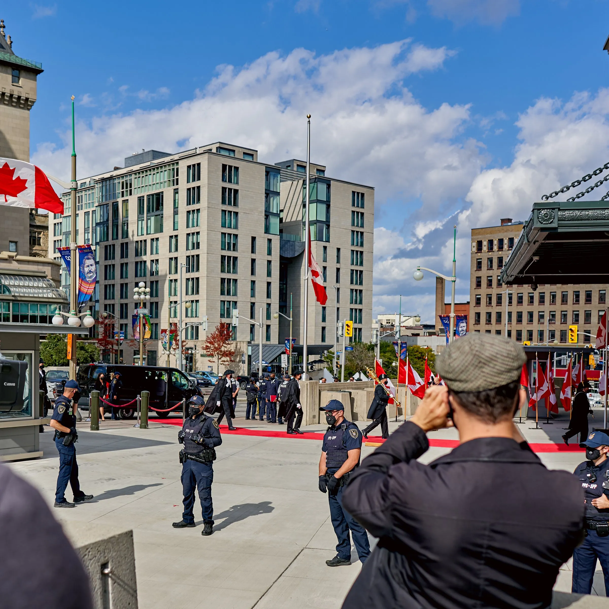 Police officers stand in a city plaza lined with Canadian flags as a man photographs them.