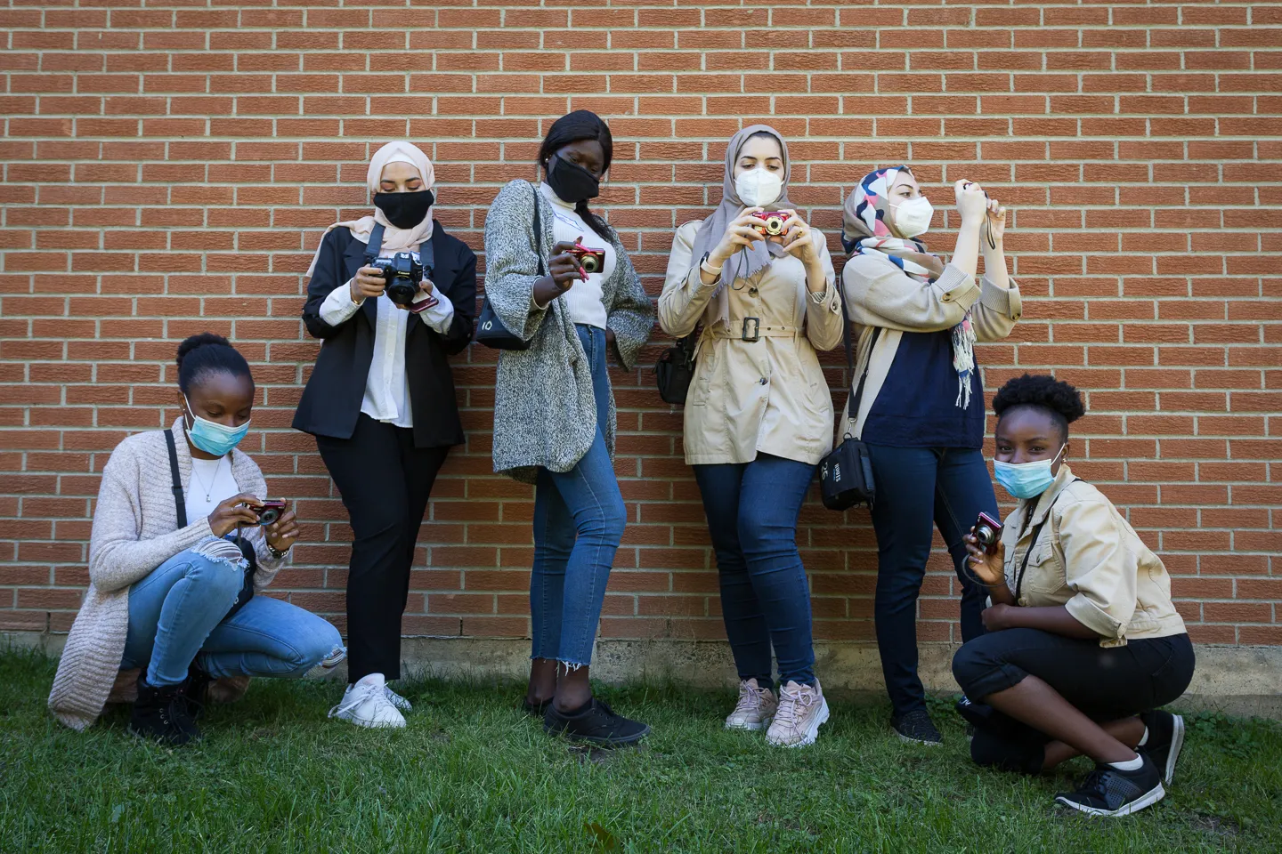 Six masked women hold cameras while standing or crouching on grass in front of a brick wall.