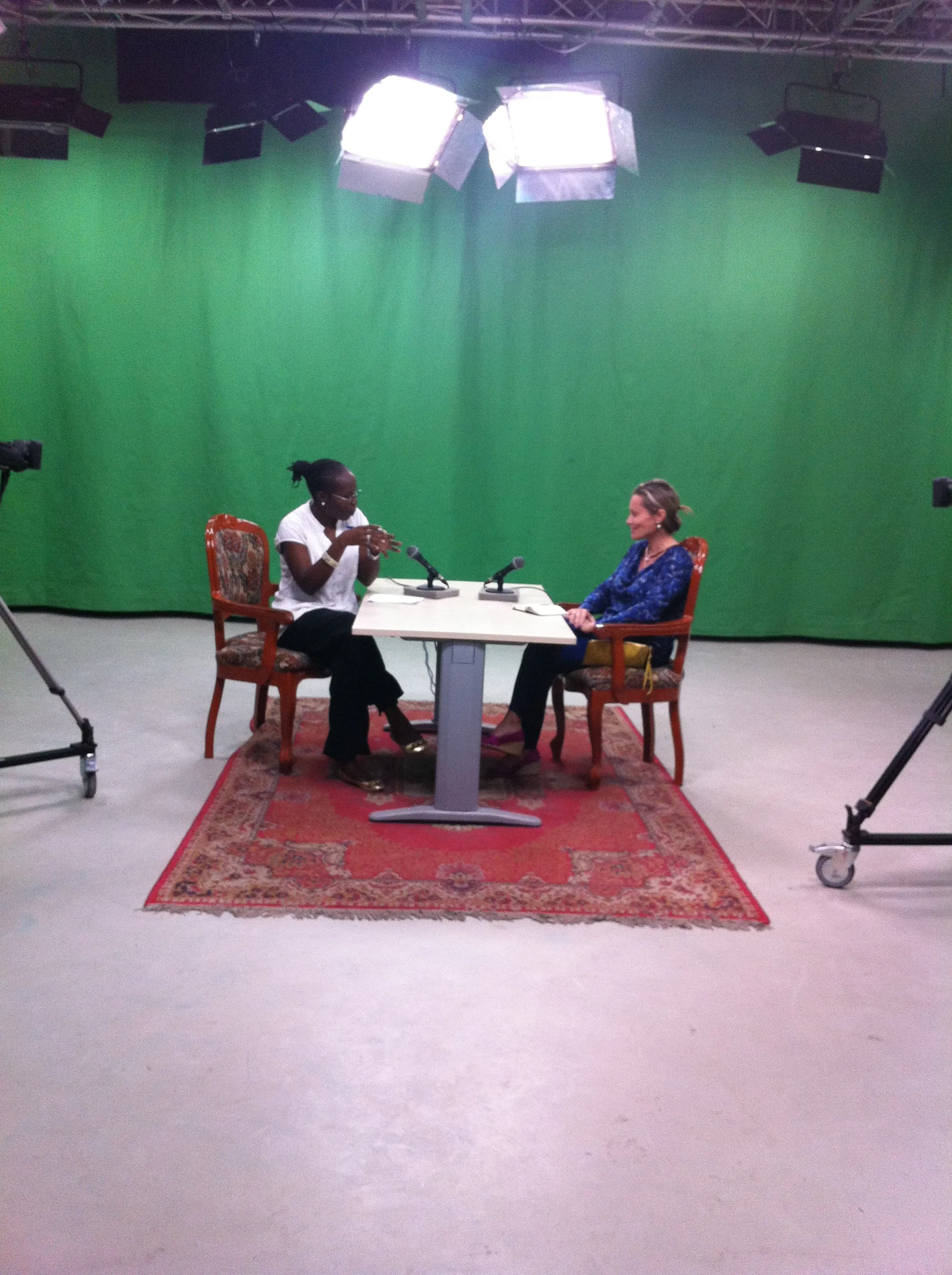 Two people sit at a table on a rug in front of a green screen, with studio lights nearby.